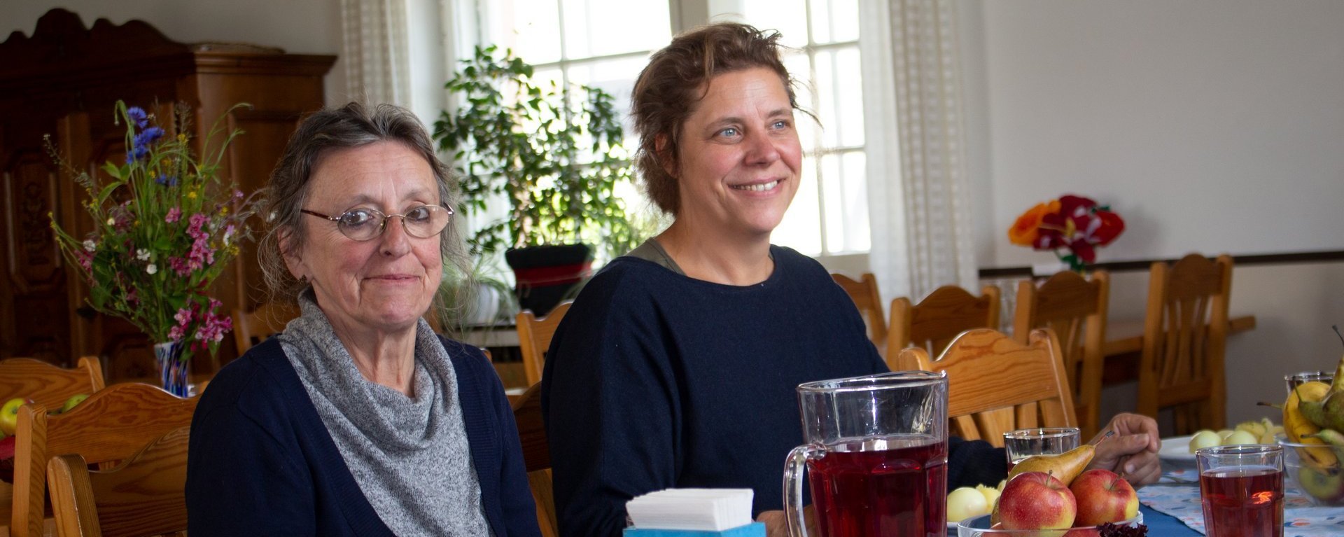 Mittagessen: zwei Frauen zu Tisch lächeln in die Kamera oder unterhalten sich. Im Vordergrund ein großer Krug Saft - es gibt Kartoffeln und Salat als Beilage - die Hauptspeise ist nicht zu sehen