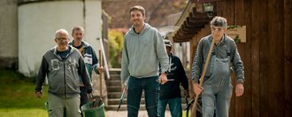 Eine Gruppe von fünf Männer laufen einen schmalen Kiesweg hinunter und tragen ihr Werkzeug für die Gartenarbeit mit sich.