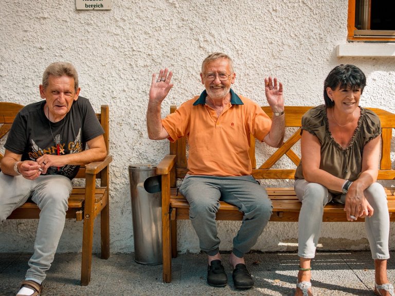 Drei Bewohnende sitzen an einer Hauswand auf einer Bank und lachen, einer von ihnen winkt fröhlich in die Kamera.