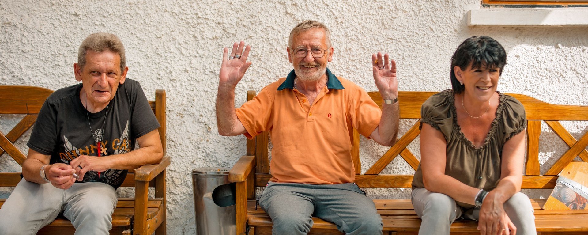 Drei Bewohnende sitzen an einer Hauswand auf einer Bank und lachen, einer von ihnen winkt fröhlich in die Kamera.