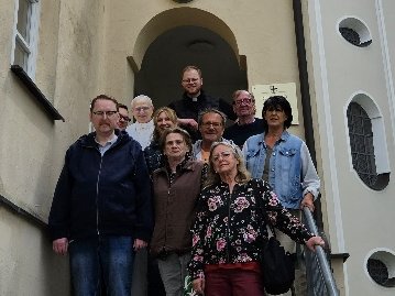 Eine Gruppe von acht Personen posiert auf einer Steintreppe vor einem weißen Kirchengebäude mit einem markanten Turm im Hintergrund.