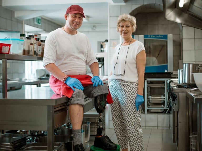 Zwei Mitarbeitenden aus der Hauswirtschaft sitzen und stehen in der Küche und lächeln.