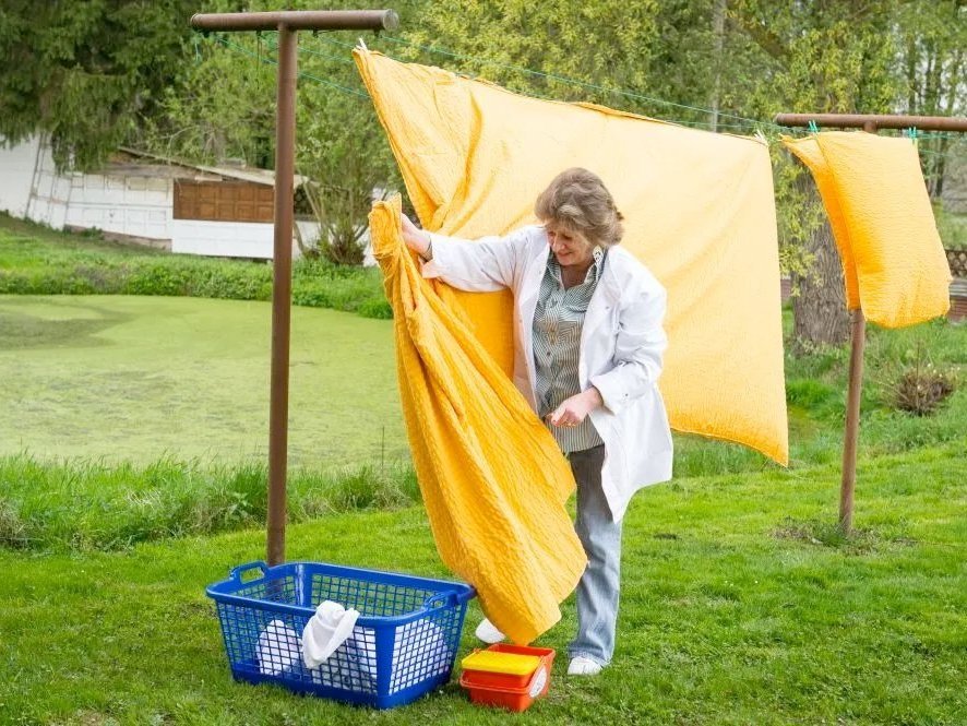 Frau mit blauem Wäschekorb beim Aufhängen gelber Bettwäsche im Garten 