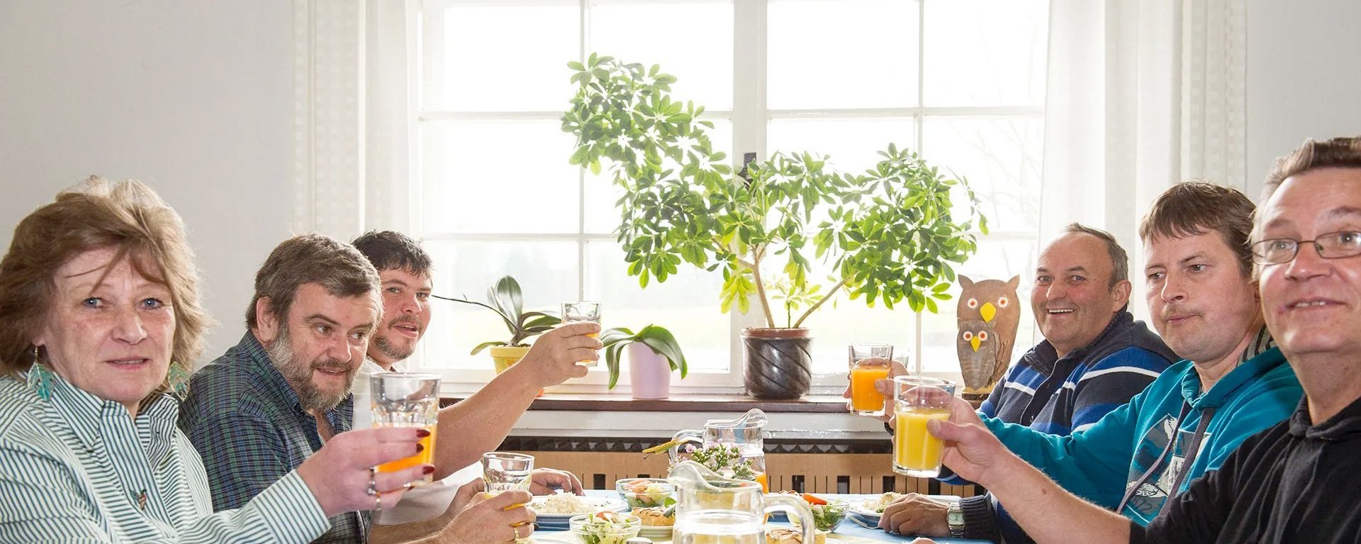 Mittagessen: Eine Gruppe Frauen und Männer prosten mit Orangensaft und Wasser der Kanera zu - im Hintergrund scheint die Sonne durch ein großes Fenster