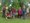 Neun Personen posieren für ein Gruppenfoto im Freien unter einem großen Baum. Mehrere tragen rote Schürzen mit einem Logo. Zwei Männer knien im Vordergrund, während die anderen sieben Personen dahinter stehen.