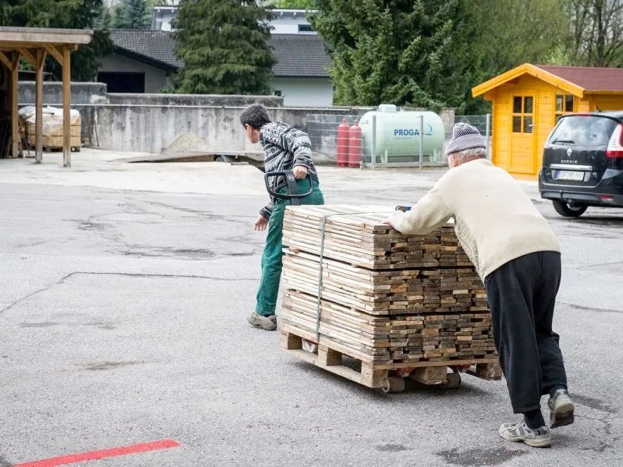 Holzlieferung: Ein Mann in grüner Arbeitshose zieht einen Hubwagen mit gestapelten Brettern. Ein zweiter, älterer Mann mit Mütze hilft schieben
