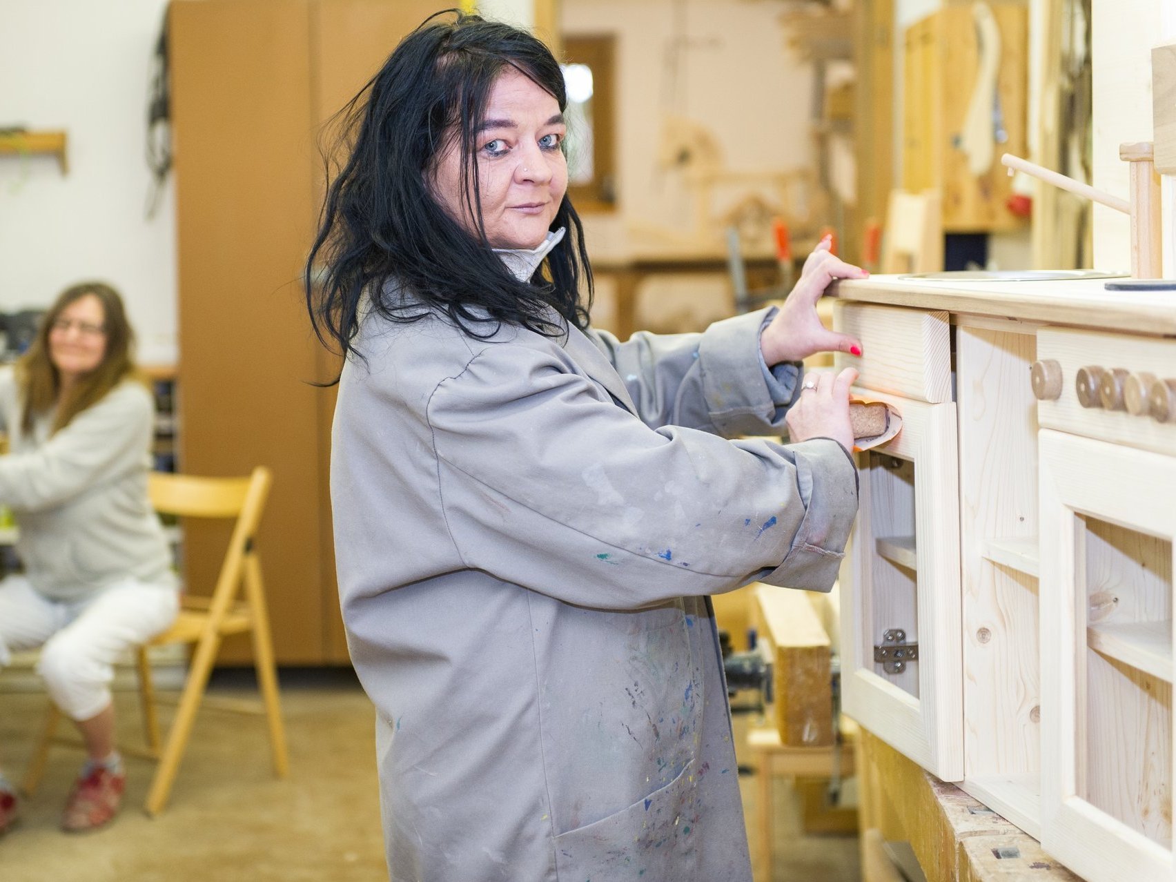 In der Holzwerkstatt: Eine Frau im grauen Arbeitskittel schmirgelt eine große Holz-Spielzeug-Küche ab.