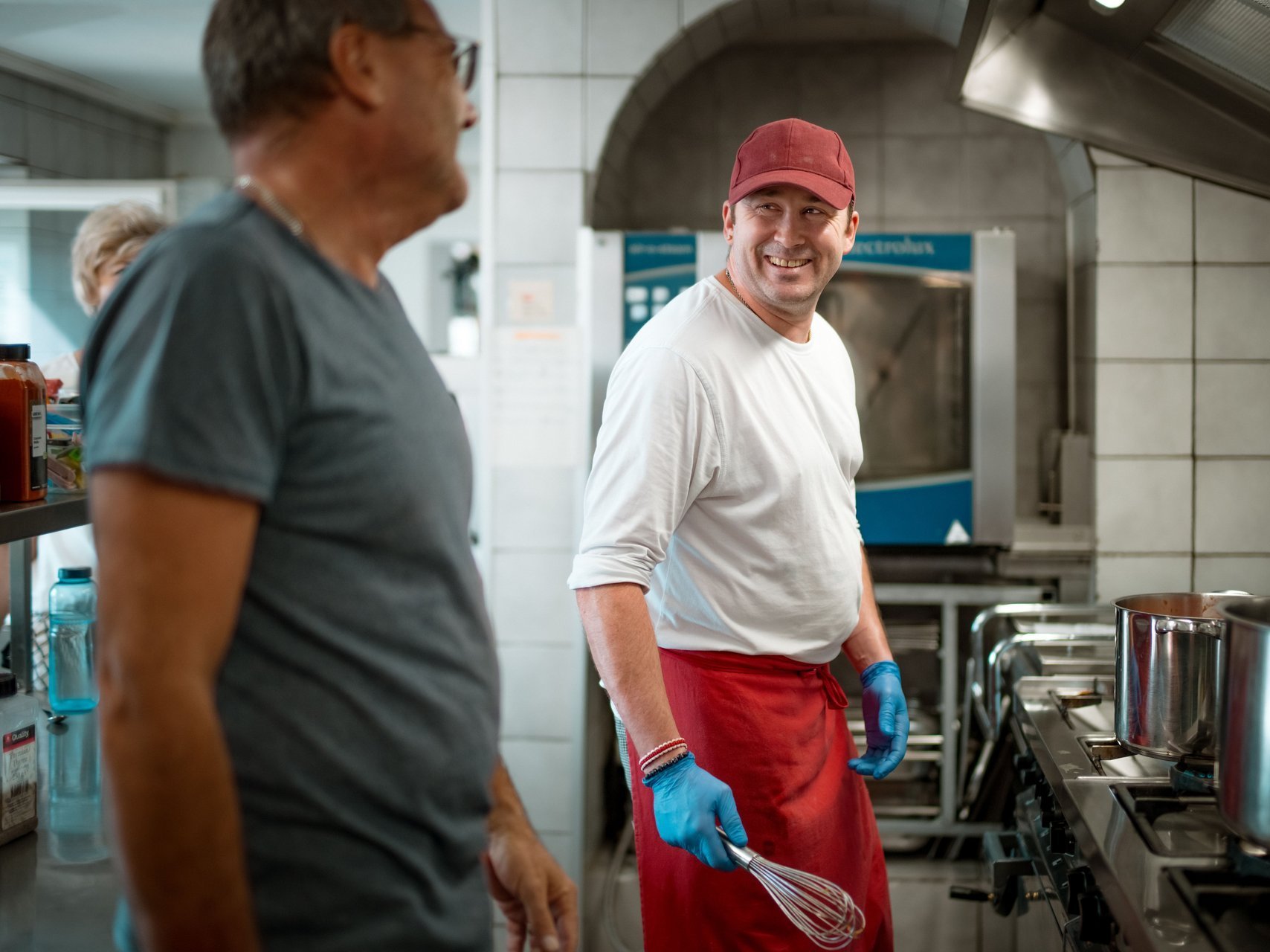 Ein Mitarbeitender und ein kochender Mitarbeitender aus der Hauswirtschaft lachen gemeinsam in der großen Küche.