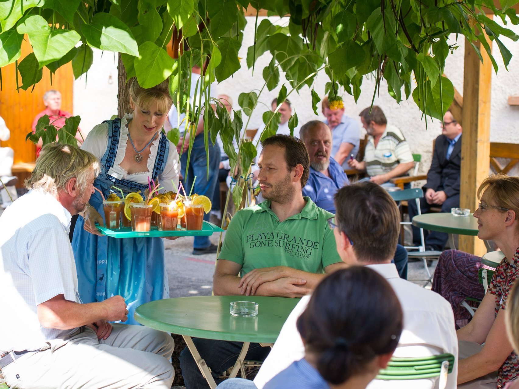 Gruppe Männer und Frauen, die im Biergarten einen Saft mit Orangenscheibe und Strohhalm von einer Kellnerin im Dirndl serviert bekommen