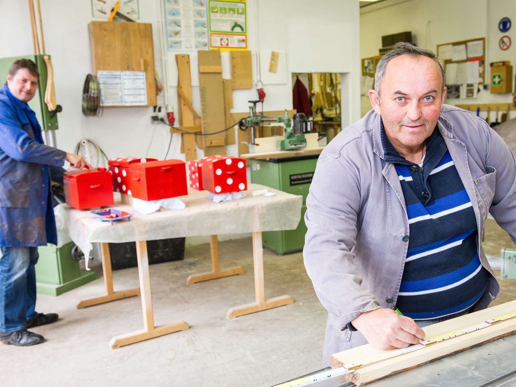 In der Holzwerkstatt ein Mann markiert die Länge eines Brettes mit dem Bleistift - im Hintergrund ein weiterer beim Lackieren von Holztruhen mit roter Farbe.