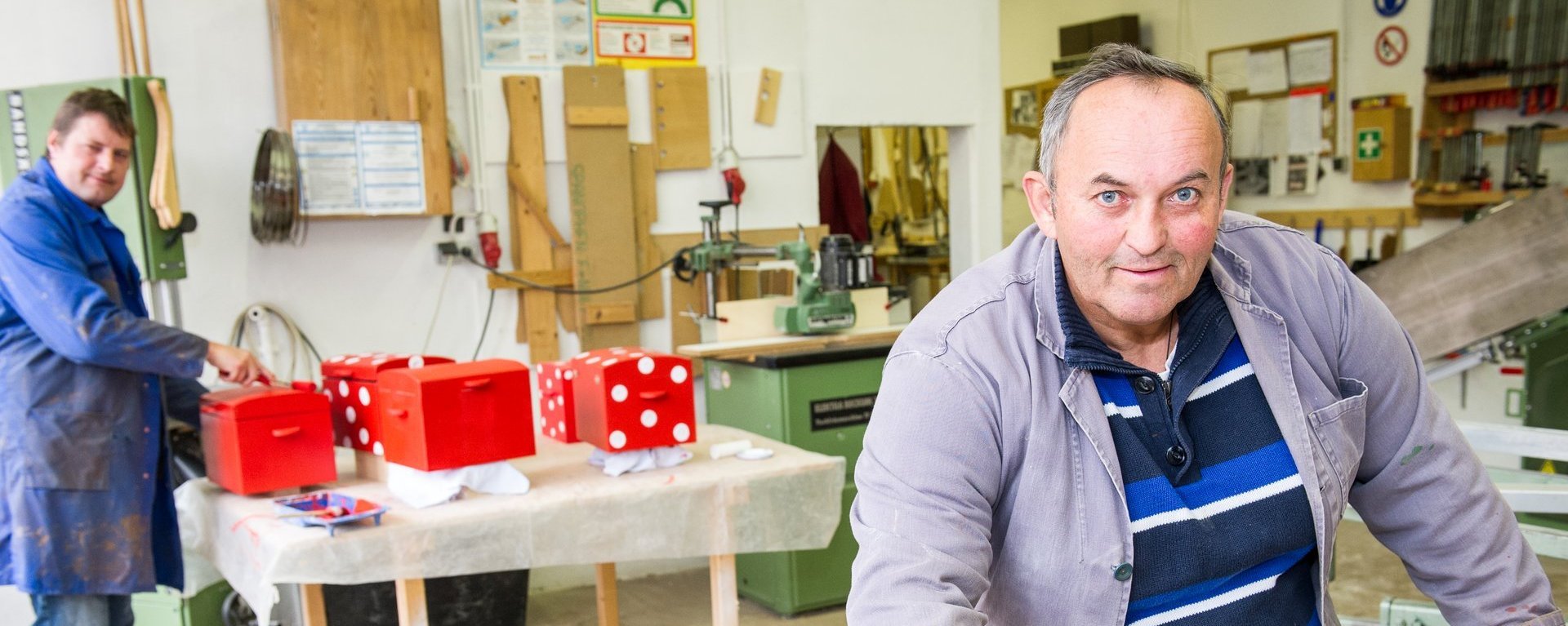 In der Holzwerkstatt ein Mann markiert die Länge eines Brettes mit dem Bleistift - im Hintergrund ein weiterer beim Lackieren von Holztruhen mit roter Farbe.