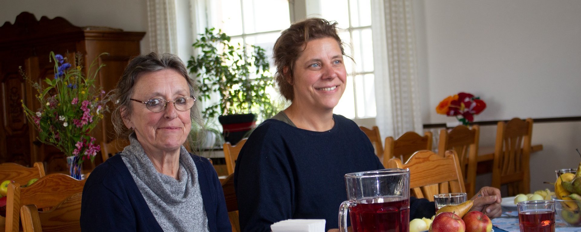 Mittagessen: zwei Frauen zu Tisch lächeln in die Kamera oder unterhalten sich. Im Vordergrund ein großer Krug Saft - es gibt Kartoffeln und Salat als Beilage - die Hauptspeise ist nicht zu sehen