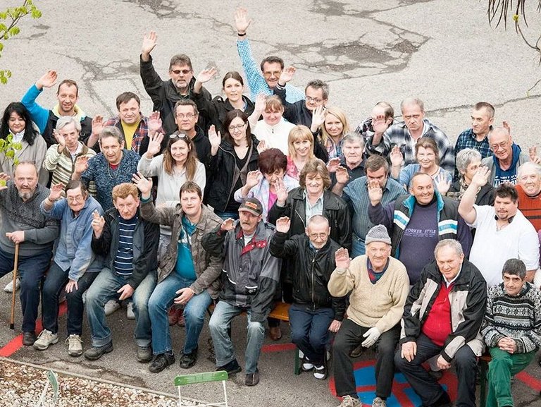 Gruppenfoto aus dem ersten Stock nach unten fotografiert zeigt ca. 25 Frauen und Männer unterschiedlichen alters, die in die Kamera winken