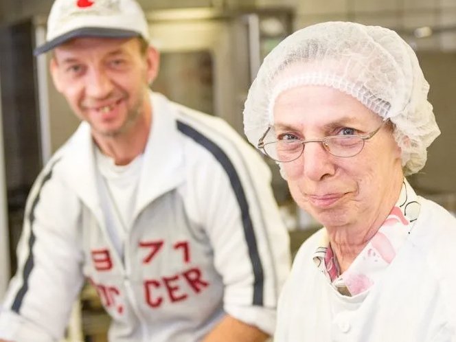 Köchin (mit Haarnetz) und ein helfender Mann hinter einer Arbeitsplatte und blicken freundlich in die Kamera