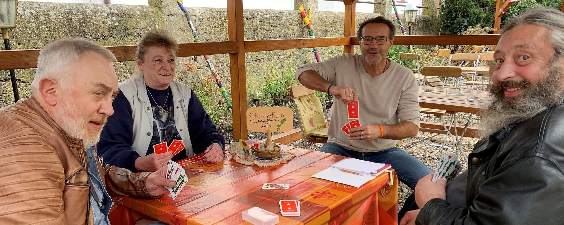 Am "Stammtisch der lustigen Schwestern und Brüder" spielen eine Frau und drei Männer Karten. Der Stammtisch wurde wohl nach draussen verlegt, um das gute Wetter zu genießen.