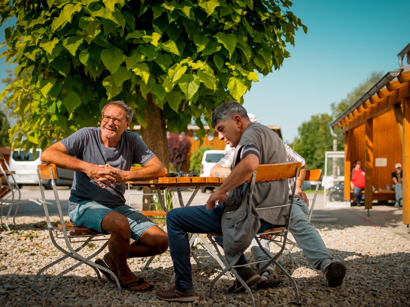 Drei Männer sitzen draußen unter einem Baum in der Sonne an einem Tisch und reden miteinander.