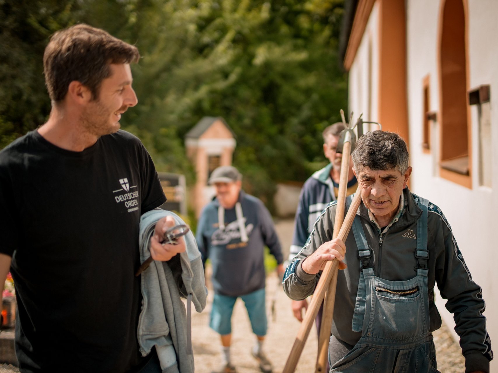 Ein Mitarbeitender und ein Bewohnender mit Gartenwerkzeug auf den Schultern, laufen einen Weg entlang.