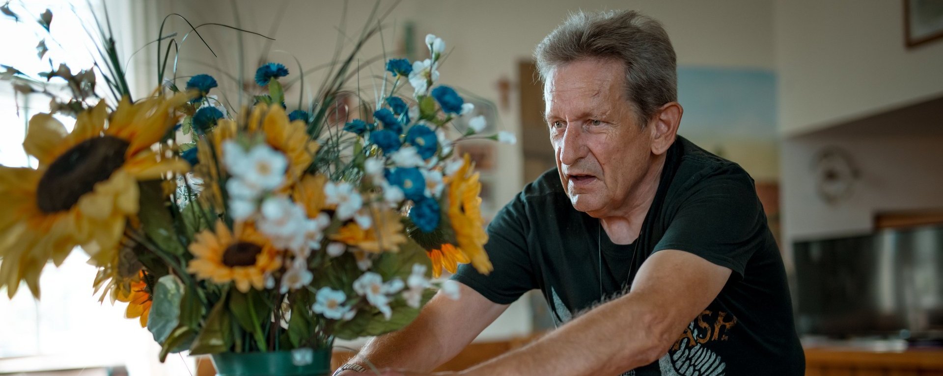 Ein Mann stellt konzentriert eine dunkelgrüne Vase mit Sonnenblumen auf einem Tisch auf.