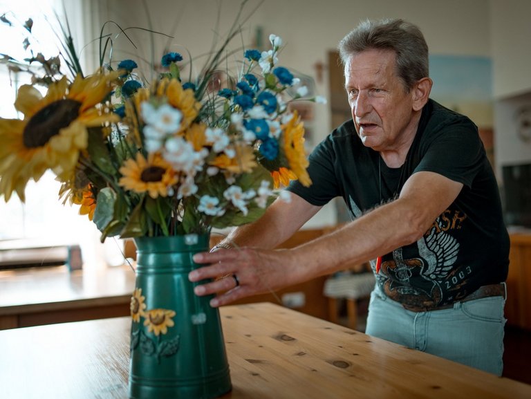 Ein Mann stellt konzentriert eine dunkelgrüne Vase mit Sonnenblumen auf einem Tisch auf.