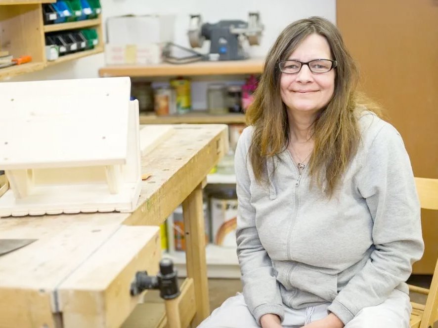 In der Schreiner-Werkstatt sitzt eine junge Frau mit Brille und langen Haaren - scheinbar stolz neben dem von ihr gefertigtem Vogelhäuschen