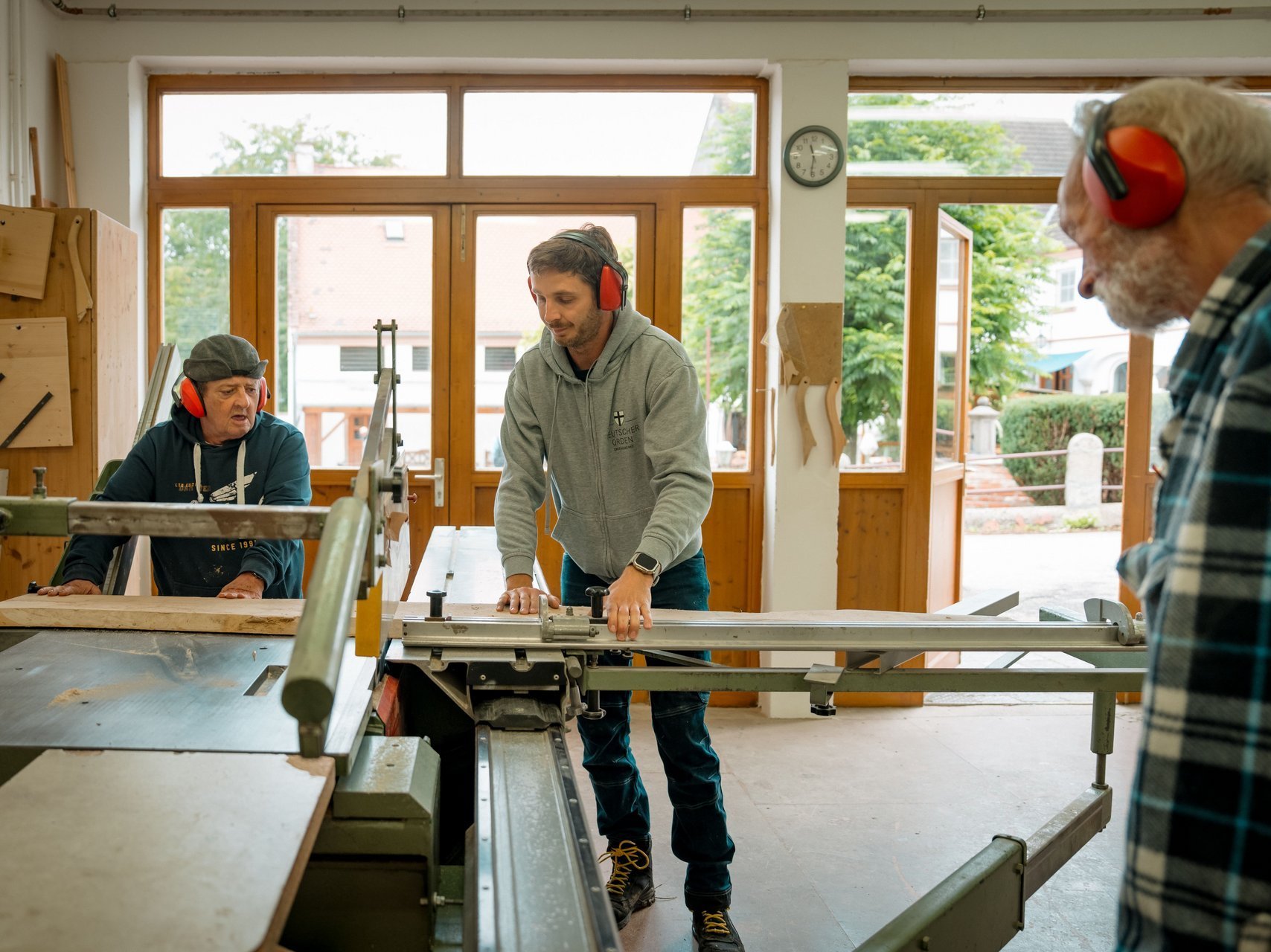 Zwei Bewohnende schneiden mit einem Mitarbeitenden in der Werkstatt ein langes Holzbrett mit einer Handkreissäge.