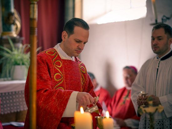 Ein Priester in roten liturgischen Gewändern gießt Flüssigkeit in einen Kelch vor brennenden Kerzen. Ein weiterer Mann in weißer Kleidung assistiert im Hintergrund.