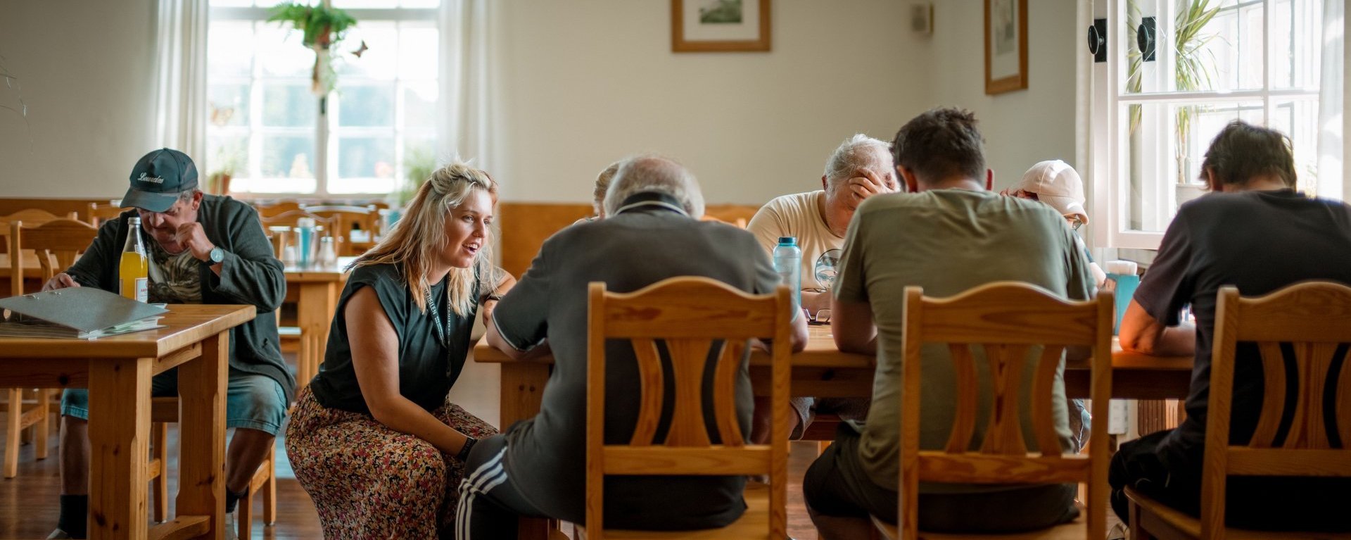 Eine Gruppe am Tisch sitzenden Bewohnenden lacht mit einer am Tisch lehnenden Mitarbeitenden.