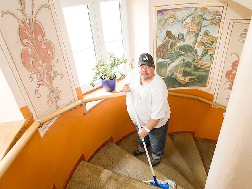 Im runden Treppenaufgang: ein Mann wischt die Stufen mit einem Wischmop - die Wände im Treppenhaus sind großflächig mit pastellfarbenen Jagdgemälden verziert 