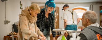 Eine Mitarbeitende steht mit einer Gruppe Bewohnenden in der Kreativwerkstatt des Hauses, diese Rollen mit viel Freude Zeitungspapier zusammen, für ein bevorstehendes Bastelprojekt.