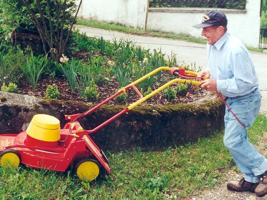 Ein älterer Mann mit Base-Cap schiebt einen knallroten Rasenmäher mit gelben Rädern knapp am Blumenbeet vorbei
