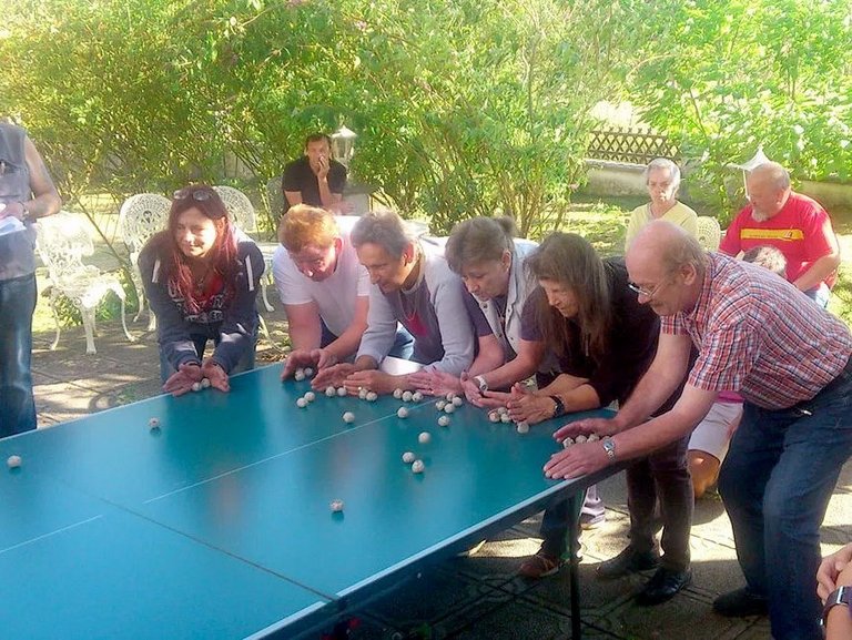 Frauen und Männer, die an einer Tischtennisplatte ein Geschicklichkeitsspiel mit Papierkugeln spielen - Punktrichter mit Schreibunterlage protokolliert