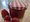 Ein Glas mit roter Marmelade und einem rot-weiß karierten Deckel steht auf einem Holztisch. Daneben liegen vier frische Erdbeeren.