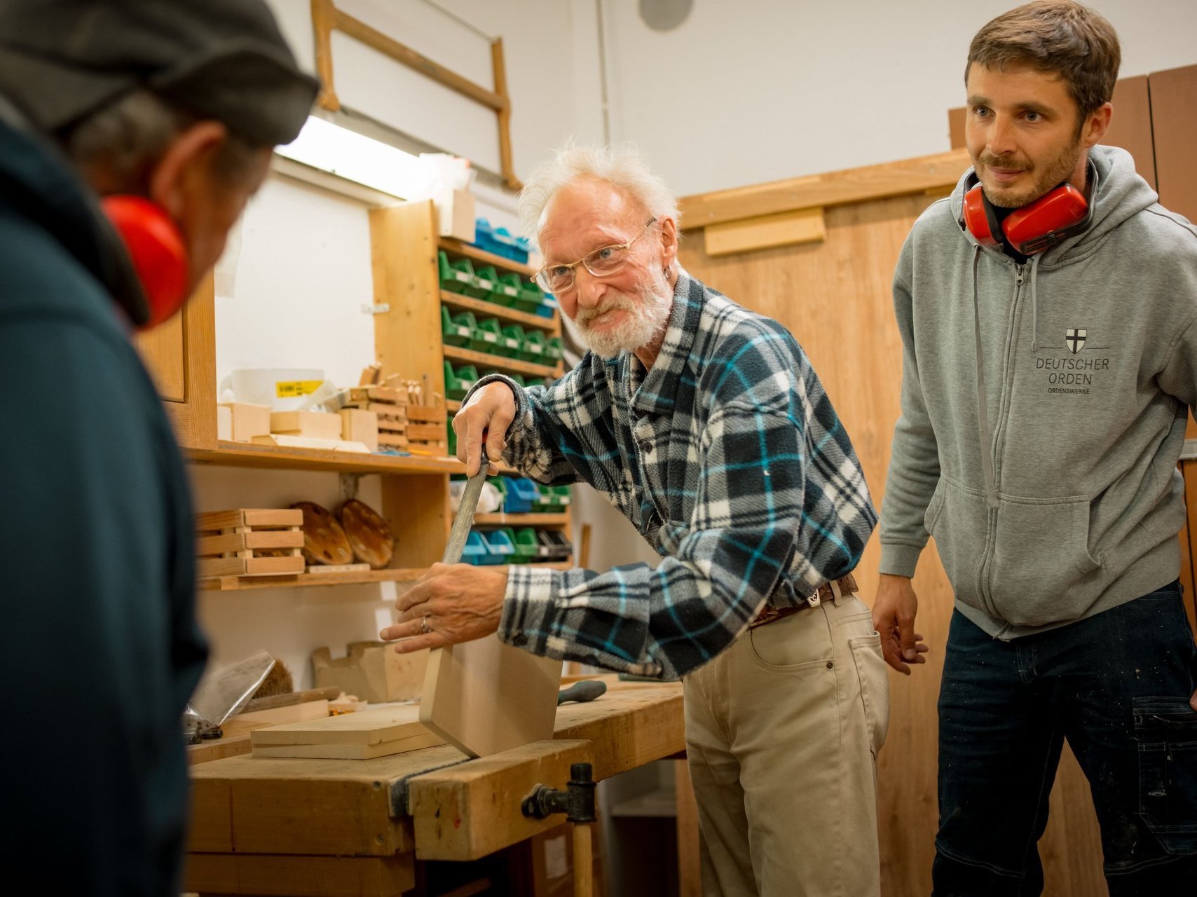 Ein Bewohnender schleift ein Stück Holz, zwei Männer schauen ihm dabei zu.. 