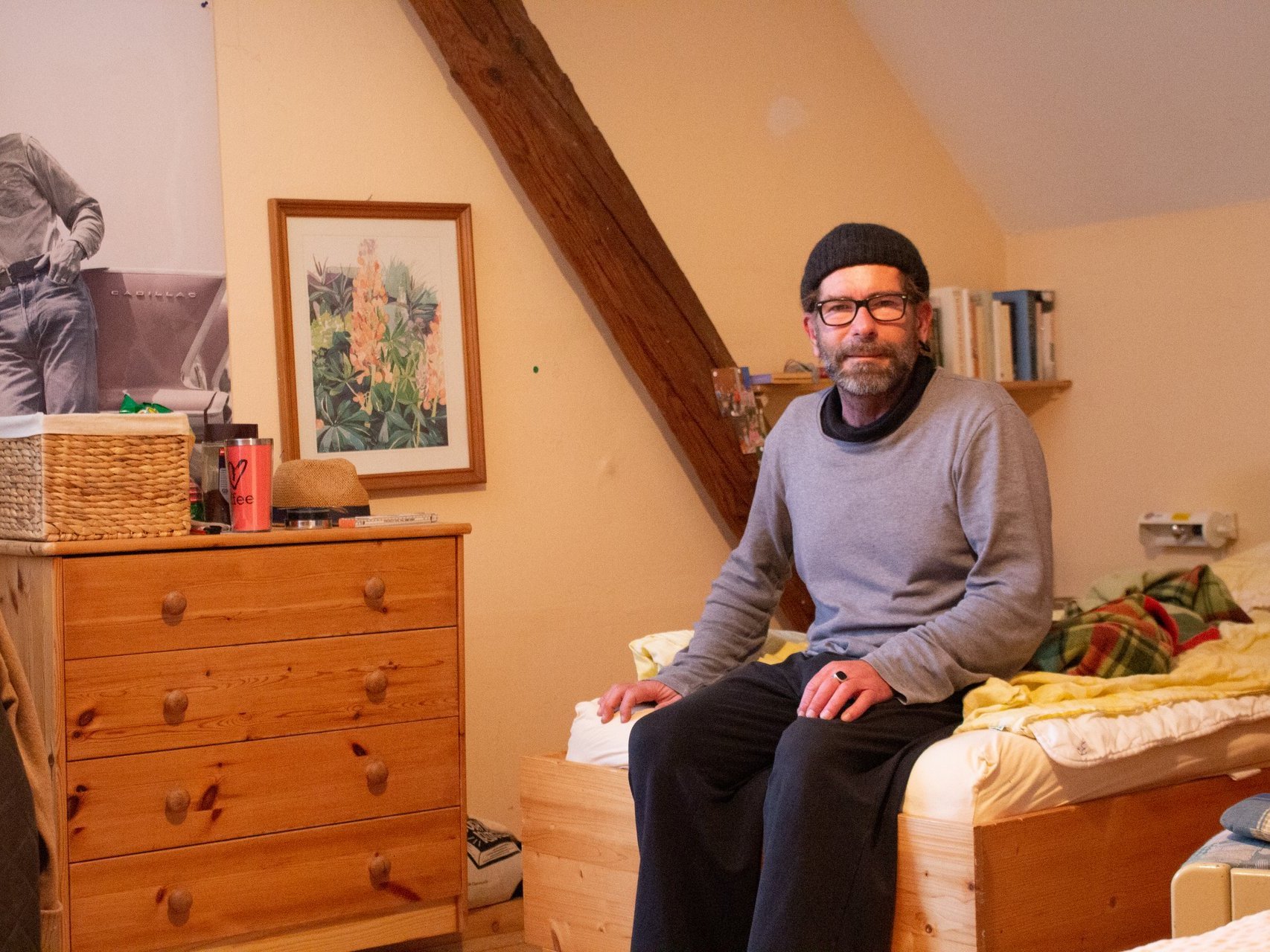 Mann mittleren alters, mit Brille, schwarzer Wollmütze und grauem Pullover sitzt auf dem Bett seines Zimmers und blickt in die Kamera. Das Zimmer sieht gemütlich aus.