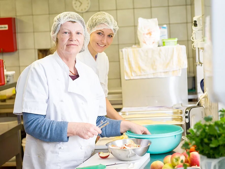 zwei Köchinnen mit Haarnetz schälen Äpfel in der Küche und lächeln in die Kamera