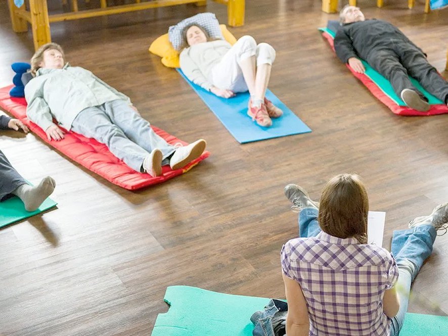 Frauen und Männer liegen auf Yoga-Matten und sehen entspannt aus - manche schlafen vielleicht schon - und machen scheinbar progressive Muskelentspannung