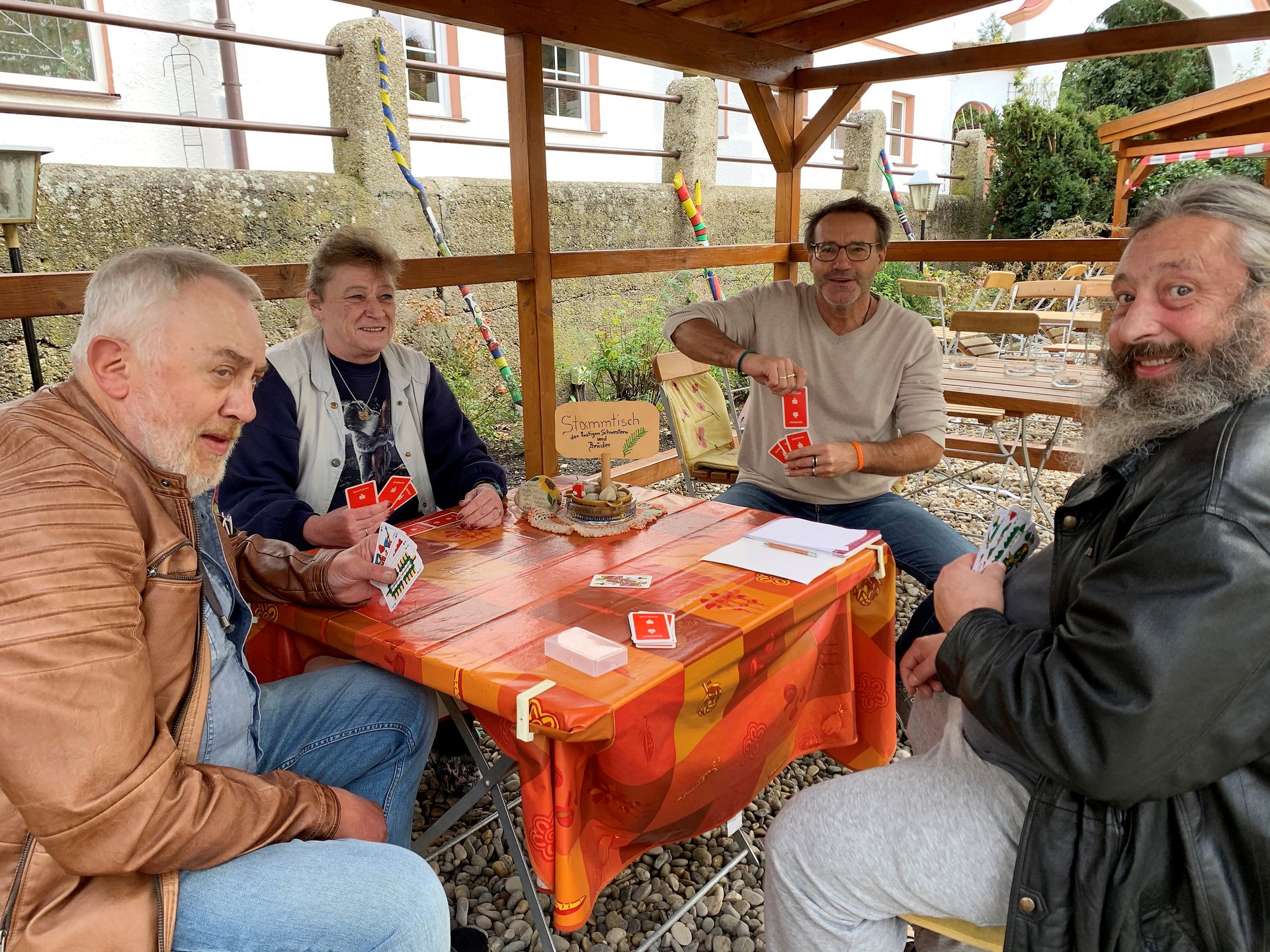 Am "Stammtisch der lustigen Schwestern und Brüder" spielen eine Frau und drei Männer Karten. Der Stammtisch wurde wohl nach draussen verlegt, um das gute Wetter zu genießen.